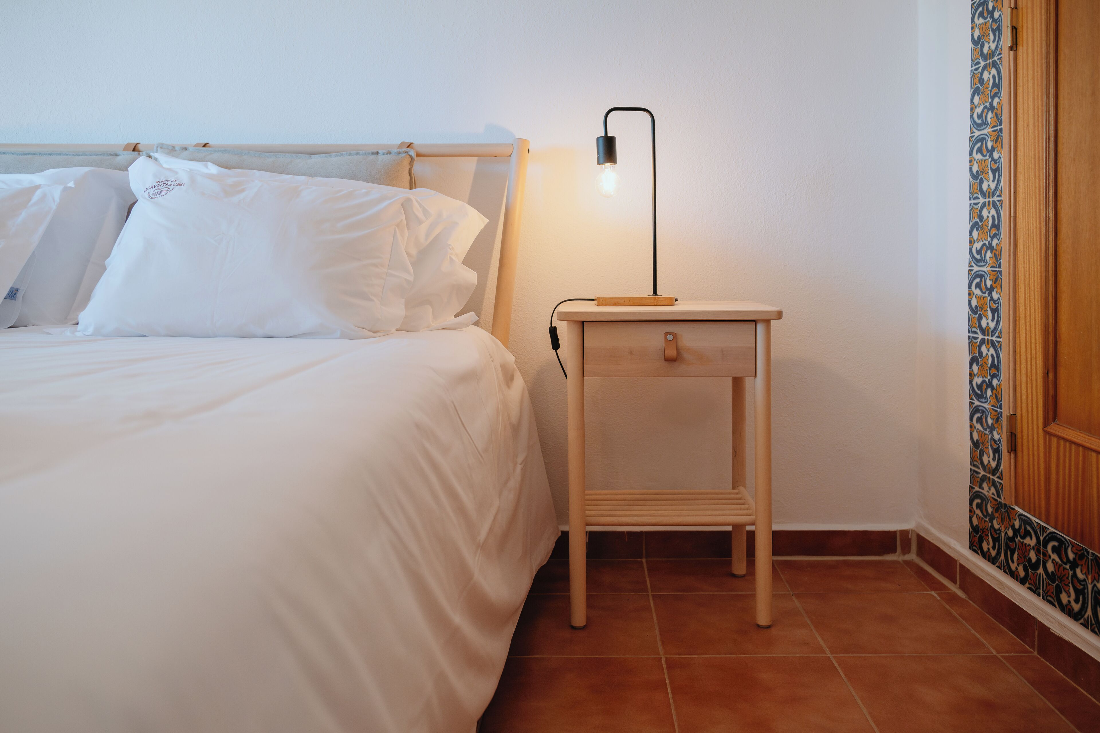 A neatly made bed with white linens and pillows, a bedside table with a lamp, and a tiled wall with a decorative border.