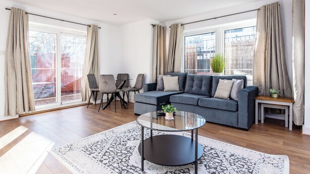 Un salon moderne avec un canapé gris, une table basse en verre et un tapis à motifs. On y trouve deux fauteuils et une petite table sur laquelle est posée une plante.