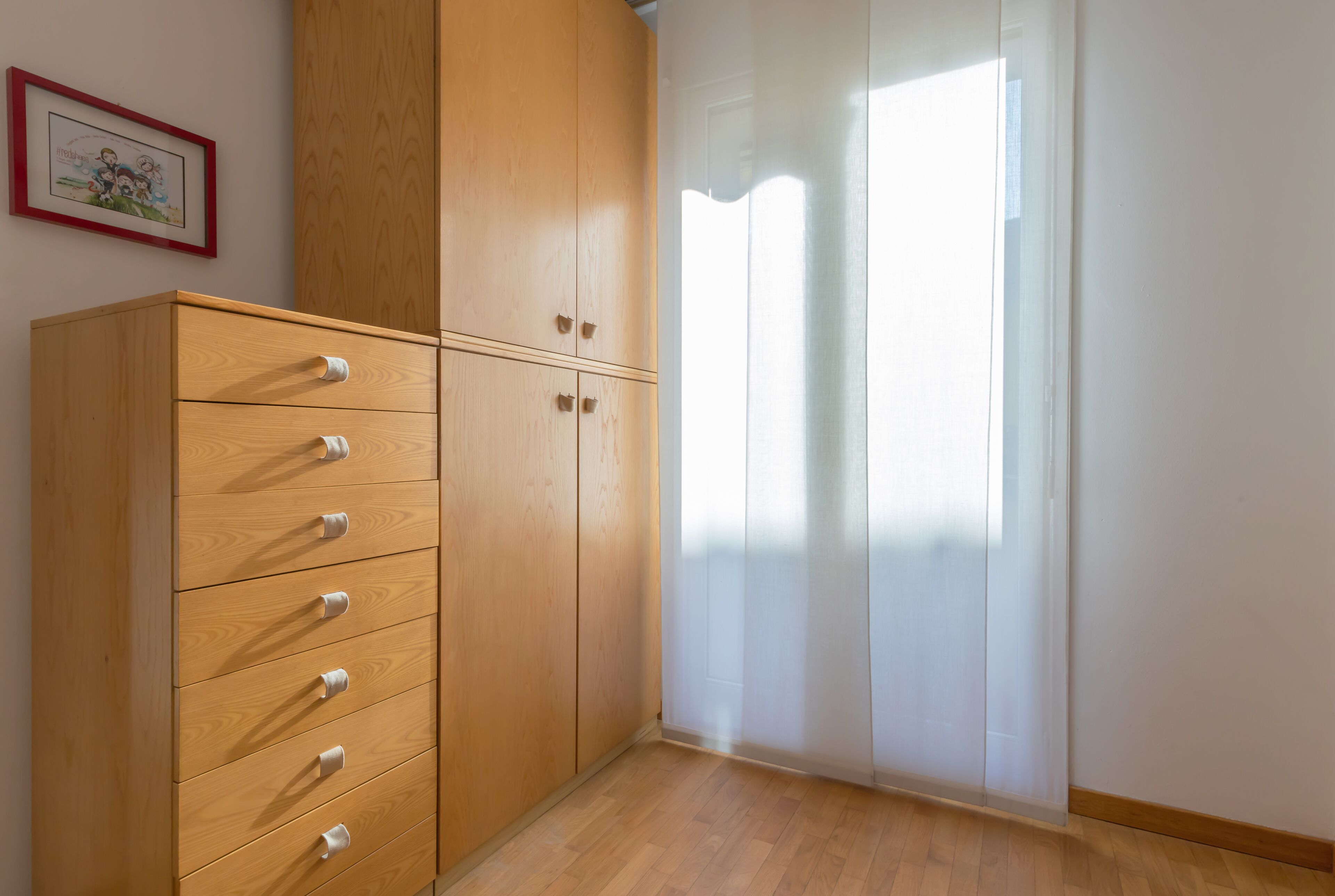 A room with a wooden wardrobe, a framed picture on the wall, and a window with sheer curtains.
