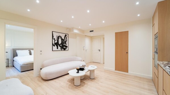 Une pièce moderne et bien éclairée, comprenant un canapé, deux petites tables, un lit et une porte donnant sur une autre pièce.