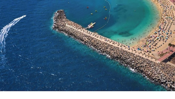 Vista aérea de una zona costera con un rompeolas rocoso, una playa con gente y un mar azul cristalino.