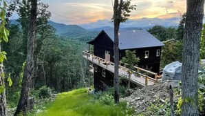 A two-story cabin with a balcony, surrounded by forested hills and a clear sky.