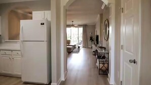 Una cocina con armarios blancos y un refrigerador, un pasillo que conduce a una sala de estar con un sofá y una mesa de centro, y una puerta a la derecha.