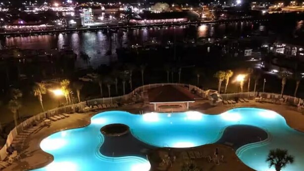 Vista aérea de un complejo de piscinas con varias albercas interconectadas, rodeado de palmeras y un paseo bien iluminado.