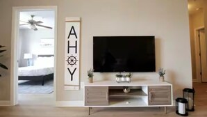 Una sala de estar moderna con un televisor de pantalla plana montado en una consola blanca, un ventilador de techo y una puerta que da a un dormitorio.