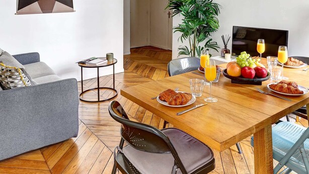 A dining area with a wooden table, chairs, and a plant. There is a sofa and a framed picture on the wall. - Charming 1BR by Pompidou & Louvre - Sleeps 4 (Paris)