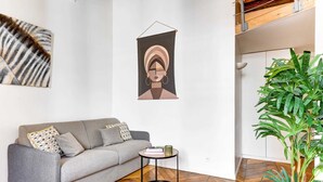 A modern living room with a grey sofa, a small round table, a hanging abstract artwork, and a potted plant. - Charming 1BR by Pompidou & Louvre - Sleeps 4 (Paris)