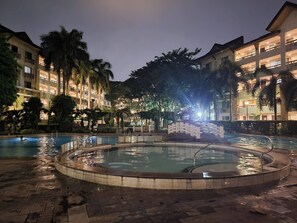 A nighttime swimming pool area with a hot tub, surrounded by palm trees and hotel buildings. - Oasis Living in Pasig City - 2bedroom Condo for Rent (Pasig)