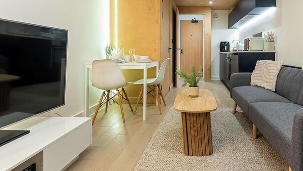 Un salon moderne avec un téléviseur à écran plat, une table à manger avec des chaises, un canapé gris, une petite table basse en bois et un coin cuisine à l’arrière-plan.