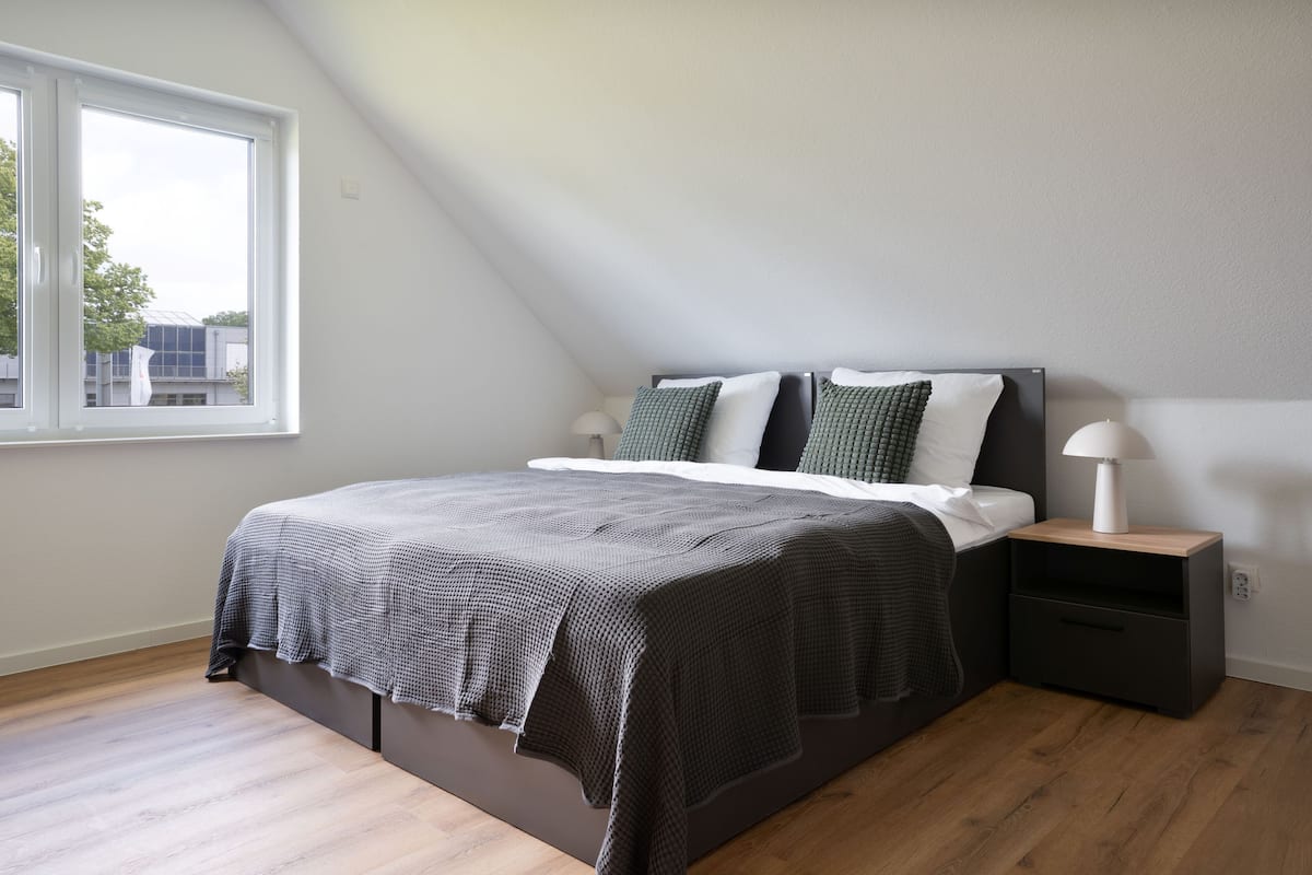 A modern bedroom with a large bed, a nightstand, a lamp, and a window with a view of buildings.