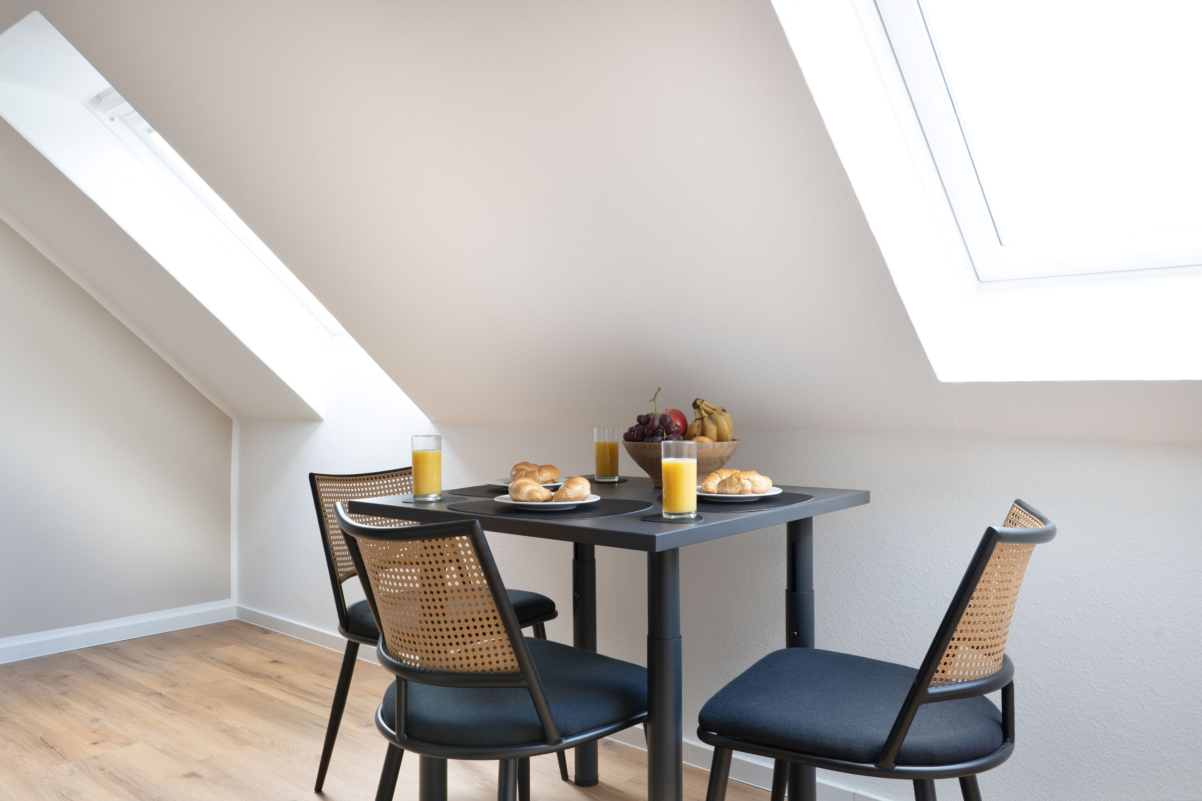 A dining area with a table set for two, featuring a fruit bowl, croissants, and glasses of orange juice.