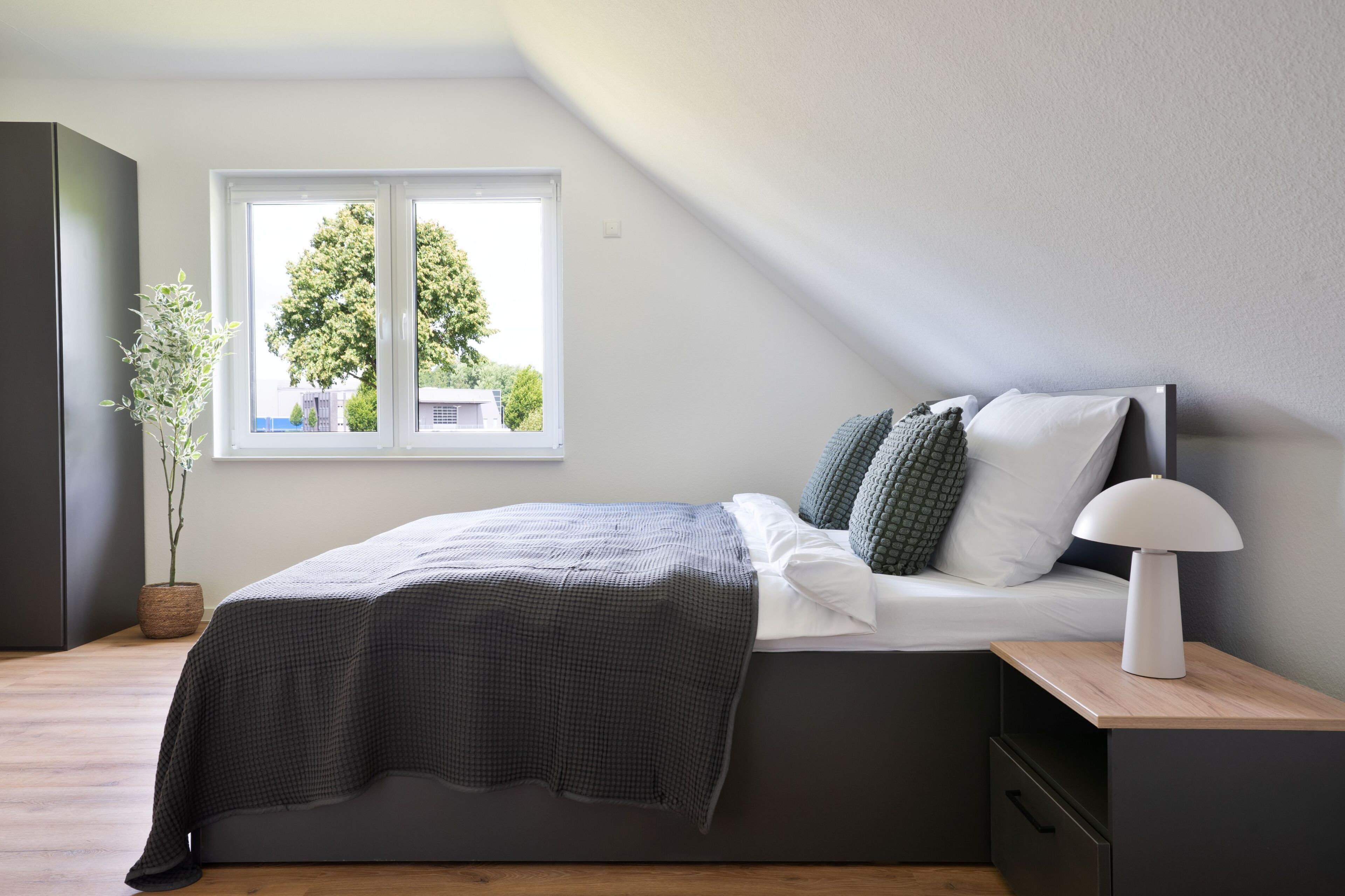Quarto moderno com cama, mesa de cabeceira, abajur e janela com vista para árvores.