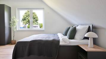 Quarto moderno com cama, mesa de cabeceira, abajur e janela com vista para árvores.