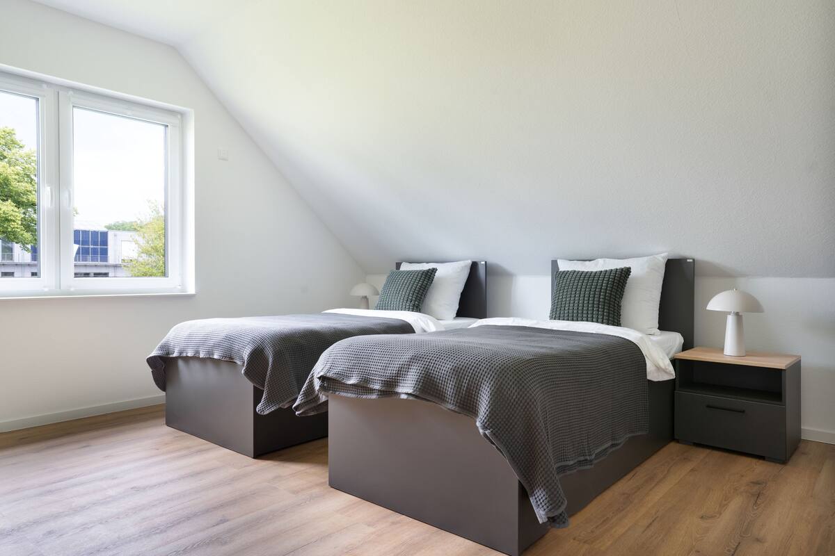 A modern bedroom with a loft bed, a bedside table with a lamp, and a large window providing natural light.