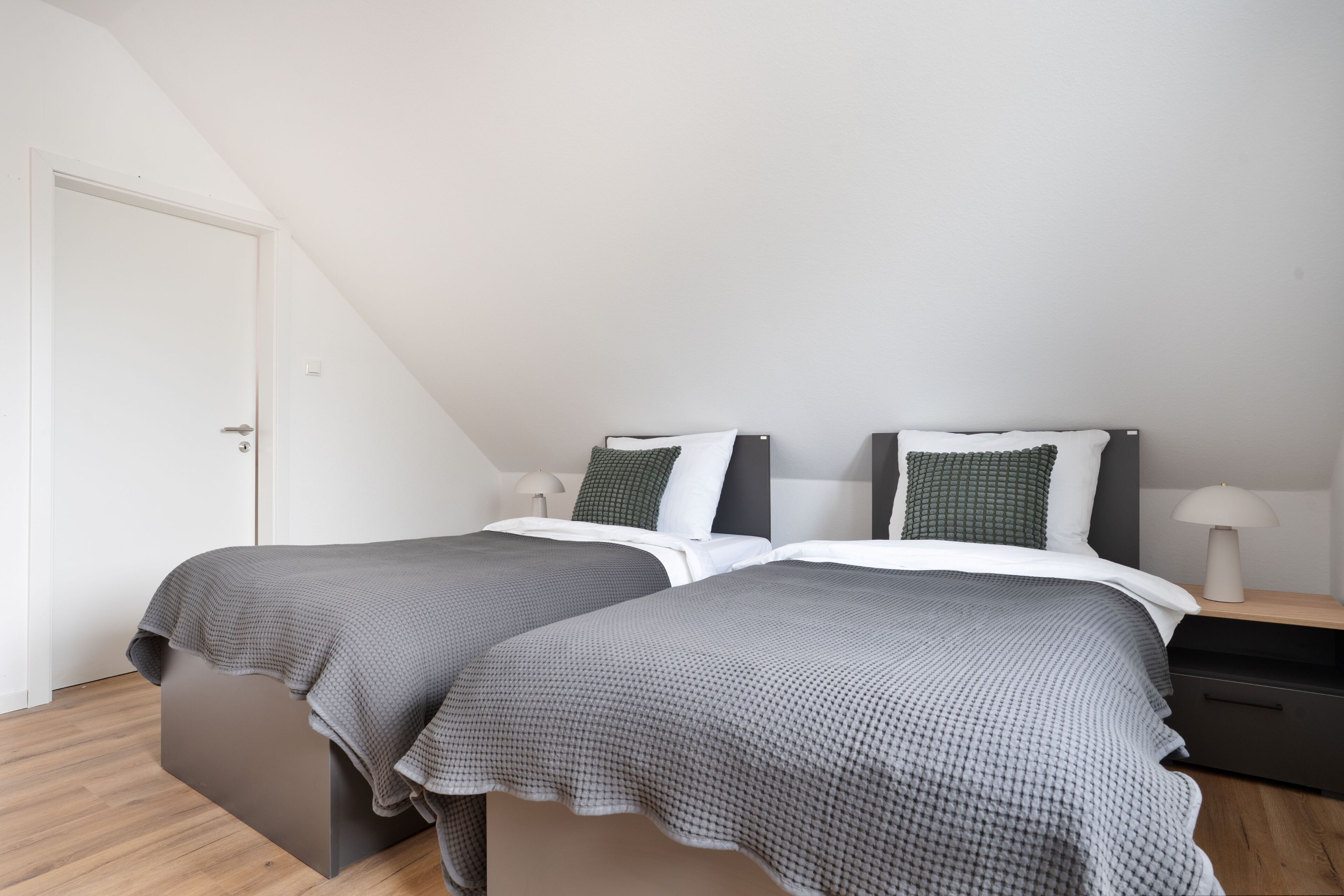A bedroom with two beds, a wooden floor, a white door, and a nightstand with a lamp.