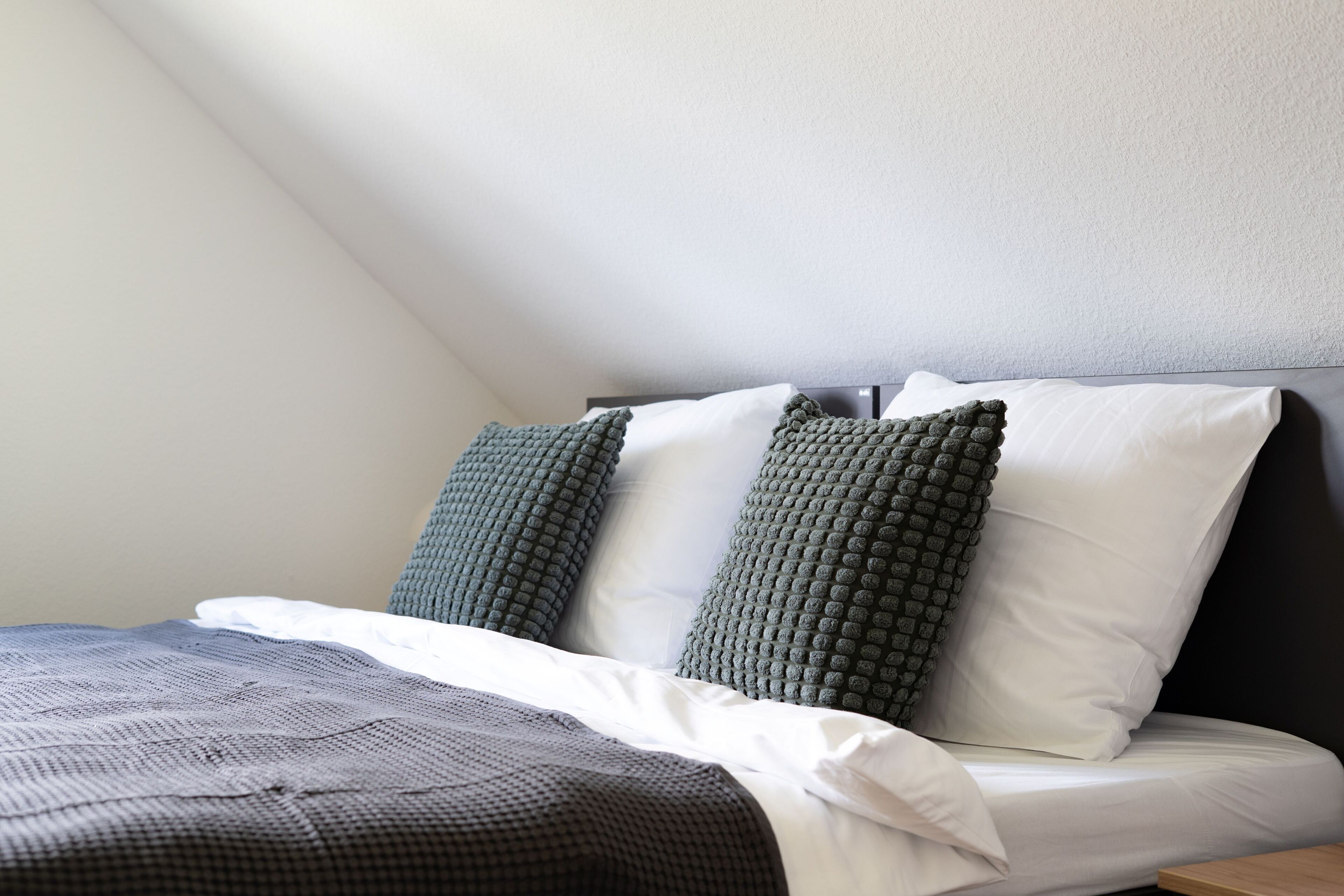 Una cama bien tendida con una colcha a cuadros y dos almohadas, una verde y una blanca, en una habitación con techo inclinado.