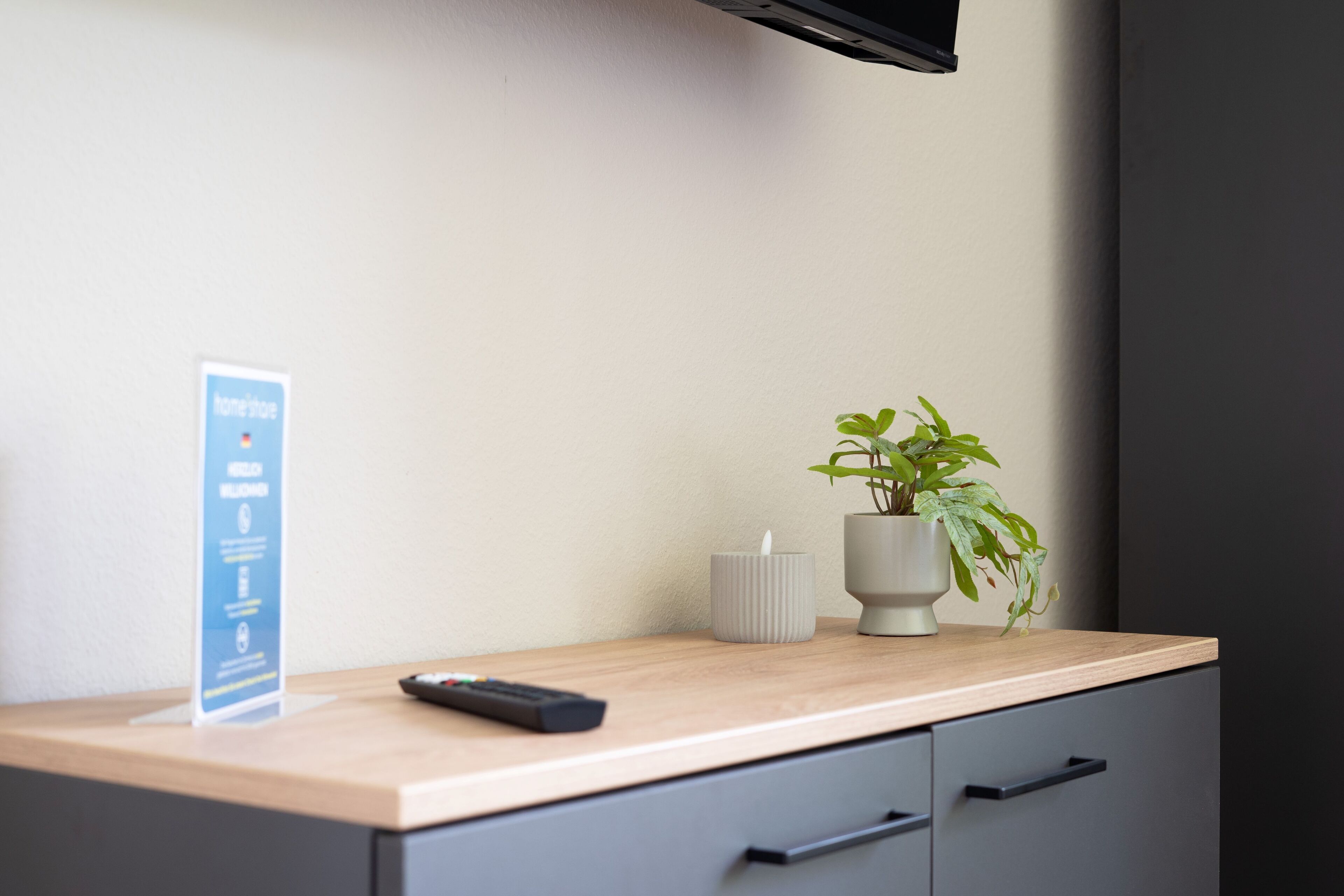 A hotel room with a wooden desk, a remote control, a potted plant, a candle, and a sign with hotel information.