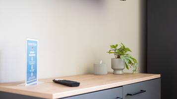 Une chambre d’hôtel avec un bureau en bois, une télécommande, une plante en pot, une bougie et un panneau d’information sur l’hôtel.