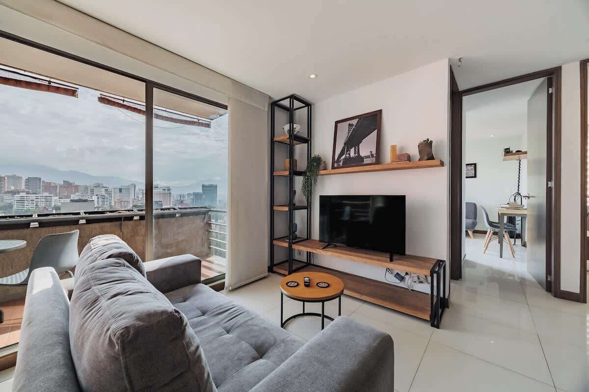A modern living room with a sofa, a TV, and a small table. There is a balcony with a city view.