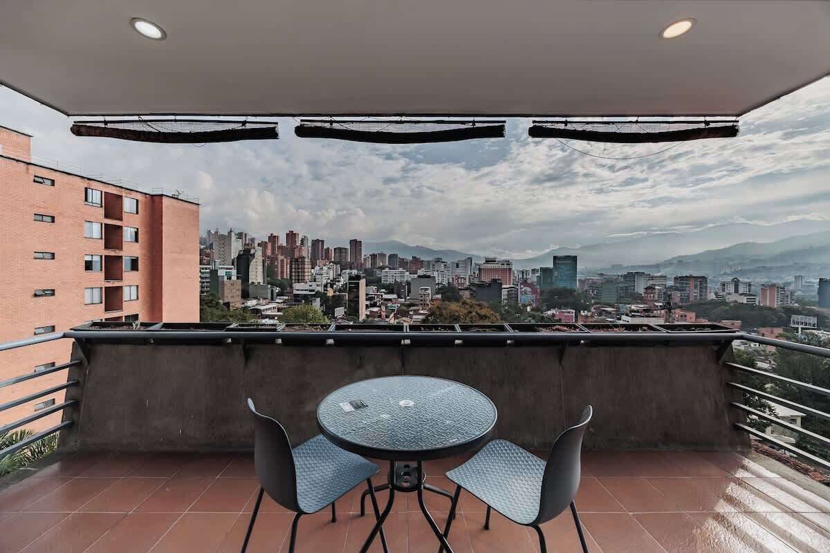 Ein Balkon mit rundem Tisch und zwei Stühlen, mit Blick auf eine Stadtlandschaft mit Gebäuden und Bergen.