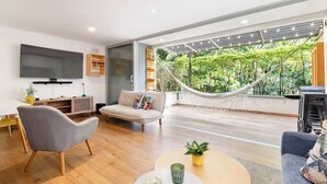 A modern living room with a large flat-screen TV, a grey sofa, a round coffee table with a plant, and a hammock overlooking a lush garden. - |EP| Bright Apt w/Terrace, AC, BBQ and Workspace (Medellín)