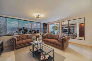 Ein Wohnzimmer mit einem großen Fenster, einem Sofa, einem Couchtisch und einem durch das Fenster einsehbar Bereich mit Küche.