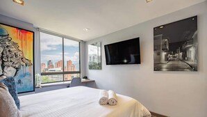 A hotel room with a large bed, a flat-screen TV mounted on the wall, and a city view through the window.