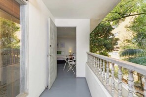 A narrow balcony with a white railing, leading to a room with a bed, a chair, and a table.