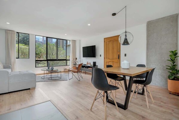 A modern living room with a dining area, large windows, a flat-screen TV, and a potted plant.