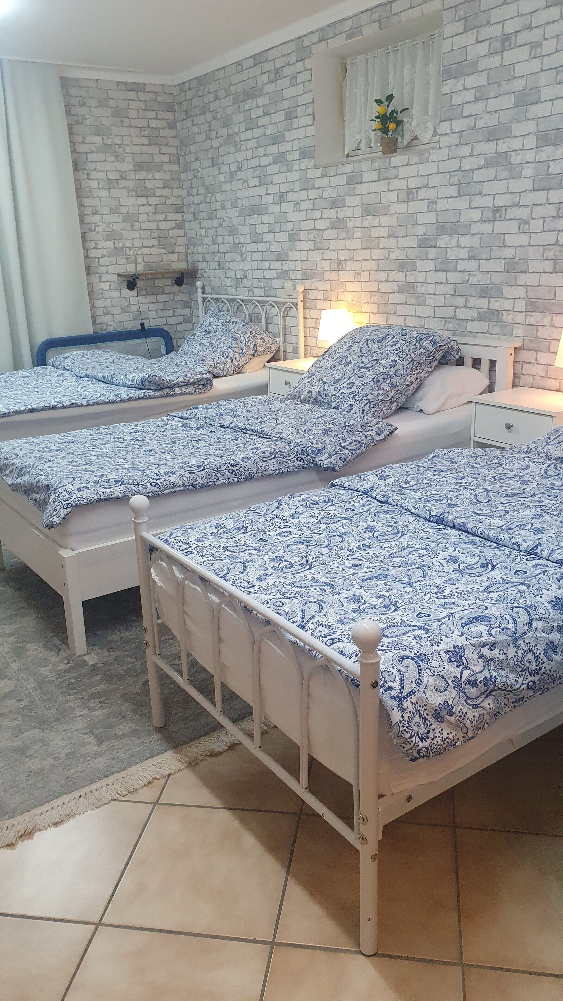 Ein Zimmer mit drei Betten, jedes mit einer gemusterten Tagesdecke, einer weißen Ziegelwand, einem kleinen Fenster mit einer Pflanze und einem Fliesenboden.