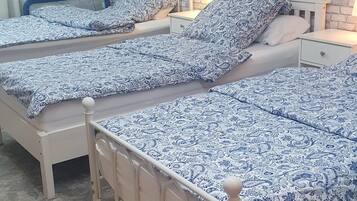 Ein Zimmer mit drei Betten, jedes mit einer gemusterten Tagesdecke, einer weißen Ziegelwand, einem kleinen Fenster mit einer Pflanze und einem Fliesenboden.