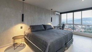 A modern bedroom with a large bed, a round side table, and a city view through large windows. - |EP| Spacious Apt with AC, Workspace, Views (Medellín)