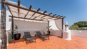 A rooftop terrace with two sun loungers, a pergola with a white canopy, potted plants, and a white wall with a decorative tower. - Retiro T2 c/Piscina em Porches por Lovelystay (Alporchinhos)