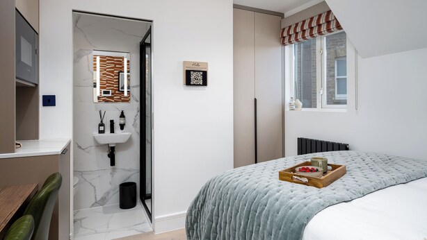 A modern bedroom with a bed, a bedside table, a window with a view, and a bathroom with a sink and mirror. - Camden | Room B3 - Studio Apartment, Sleeps 2 (London)