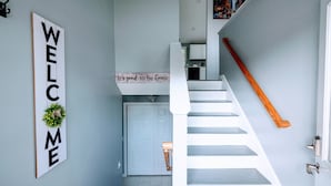 Eine Treppe mit weißer Brüstung führt zu einer Tür, einem 'WELCOME'-Schild und einem kleinen Tisch mit der Aufschrift 'es ist schön, wieder da zu sein'.