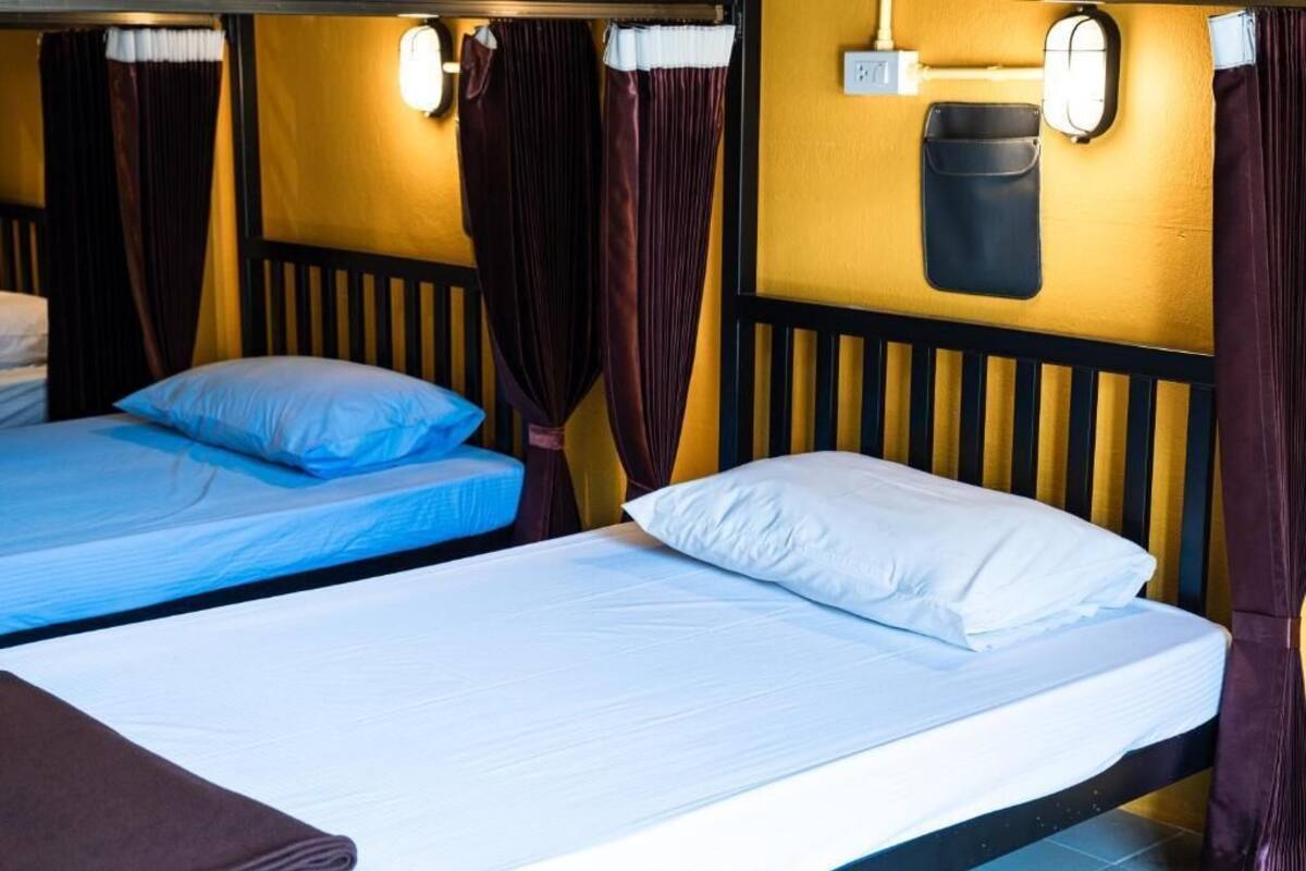 Quarto com duas camas, cada uma com roupa de cama azul e branca, e uma cabeceira preta. Há cortinas e um ponto de luz fixado na parede.