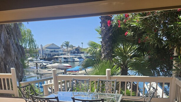 A wooden deck with a glass-top table and metal chairs, overlooking a marina with boats and palm trees. - Waterfront Elegance (Discovery Bay)