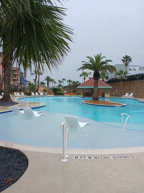A swimming pool with lounge chairs, palm trees, and a building with a red roof in the background. - Roseate Roost (Galveston)