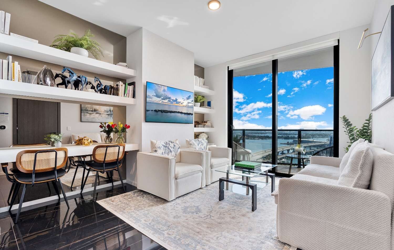 A modern living room with a dining area, large windows with a city view, and a bookshelf with decorative items.