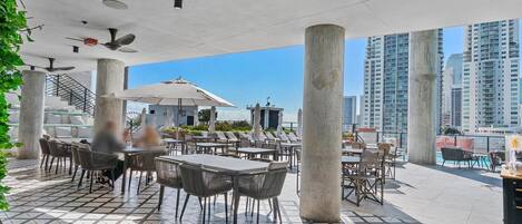 Un área de patio al aire libre con suelo de baldosas, pilares de concreto y mesas con sombrillas.