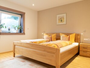 A bedroom with a wooden bed, bedside tables, a window with plants, and a painting on the wall.