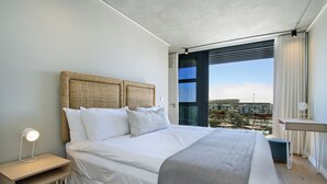 A modern bedroom with a large bed, a bedside table with a lamp, a view of the city through a large window, and a small table by the window.