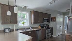 A modern kitchen with dark wooden cabinets, stainless steel appliances, and a large window providing natural light. - Coastal Trail Getaway (Port Hood)