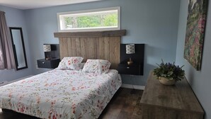 A bedroom with a wooden headboard, a large bed with floral bedding, two bedside tables with lamps, a window with a view of greenery, and a potted plant on a wooden bench. - Coastal Trail Getaway (Port Hood)