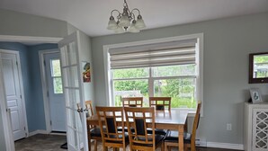 A dining area with a table and chairs, a large window with blinds, and a chandelier. - Coastal Trail Getaway (Port Hood)