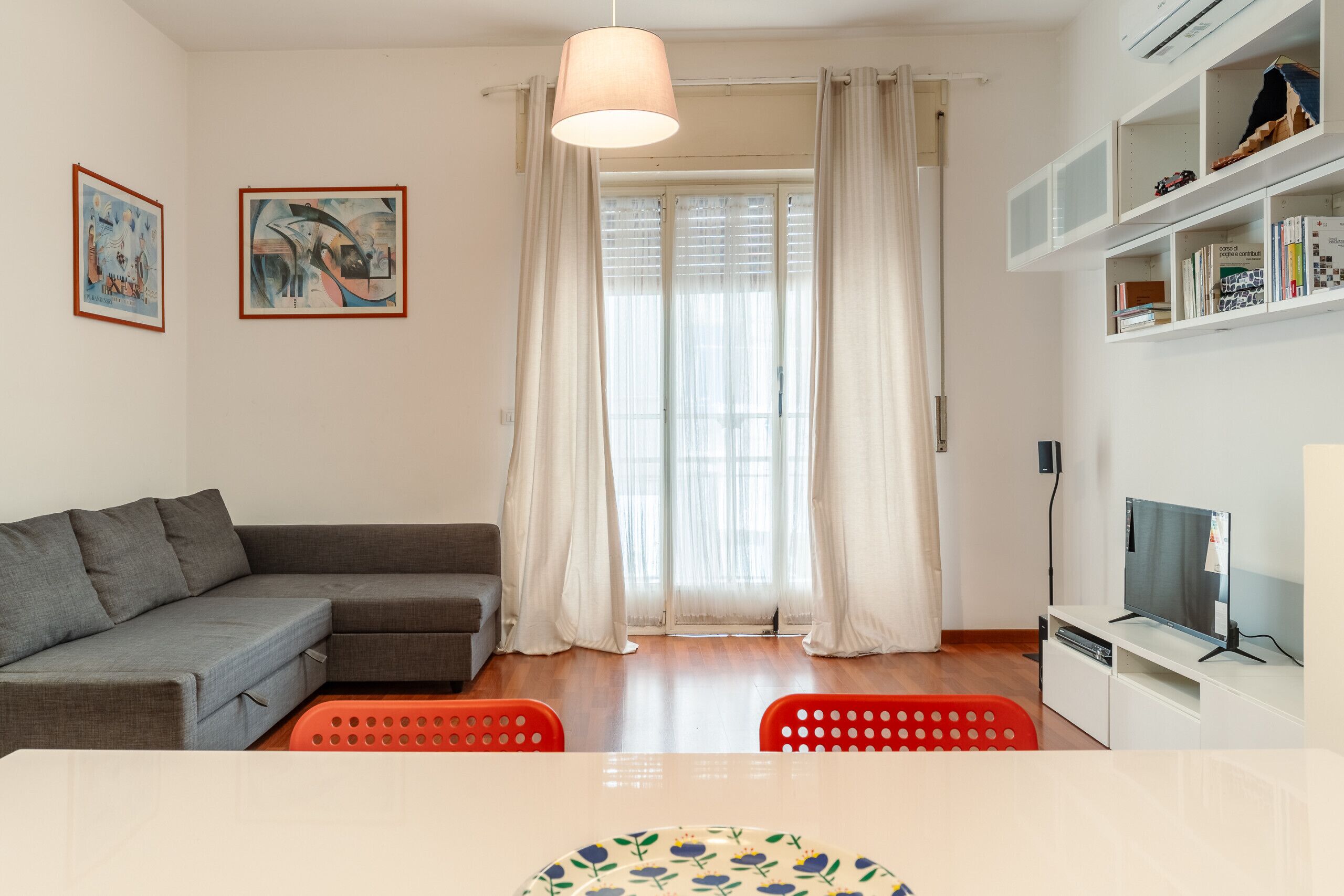 Una sala de estar moderna con un sofá gris, una mesa de comedor con mantel floreado y un televisor sobre un mueble blanco.