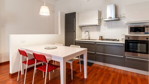 A modern kitchen with white cabinetry, stainless steel appliances, and a dining area with a white table and red chairs. - Bacchilide Novanta by Wonderful Italy (Syracuse)