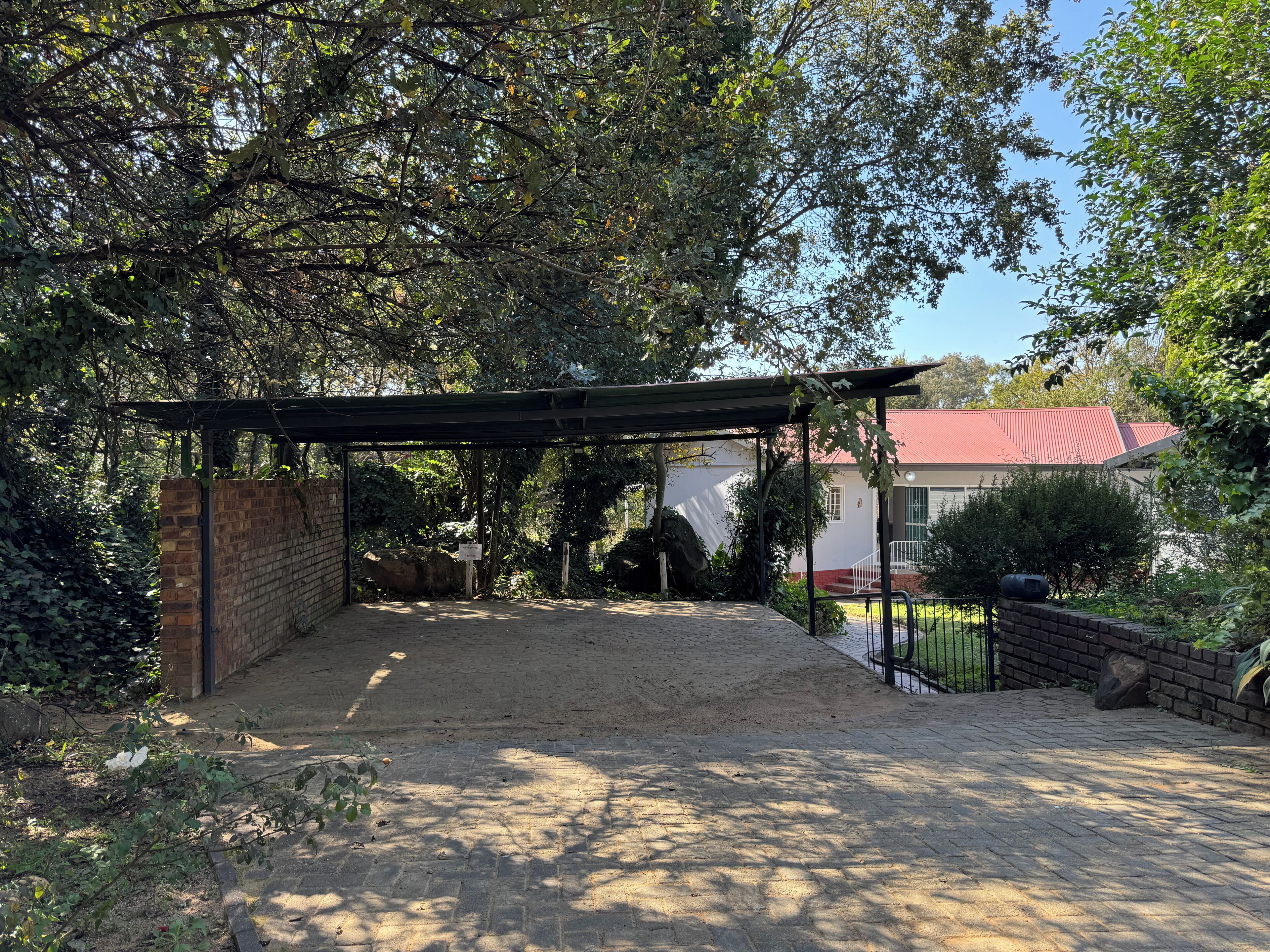 A covered outdoor area with a brick wall, a paved path, and a red-roofed building in the background.