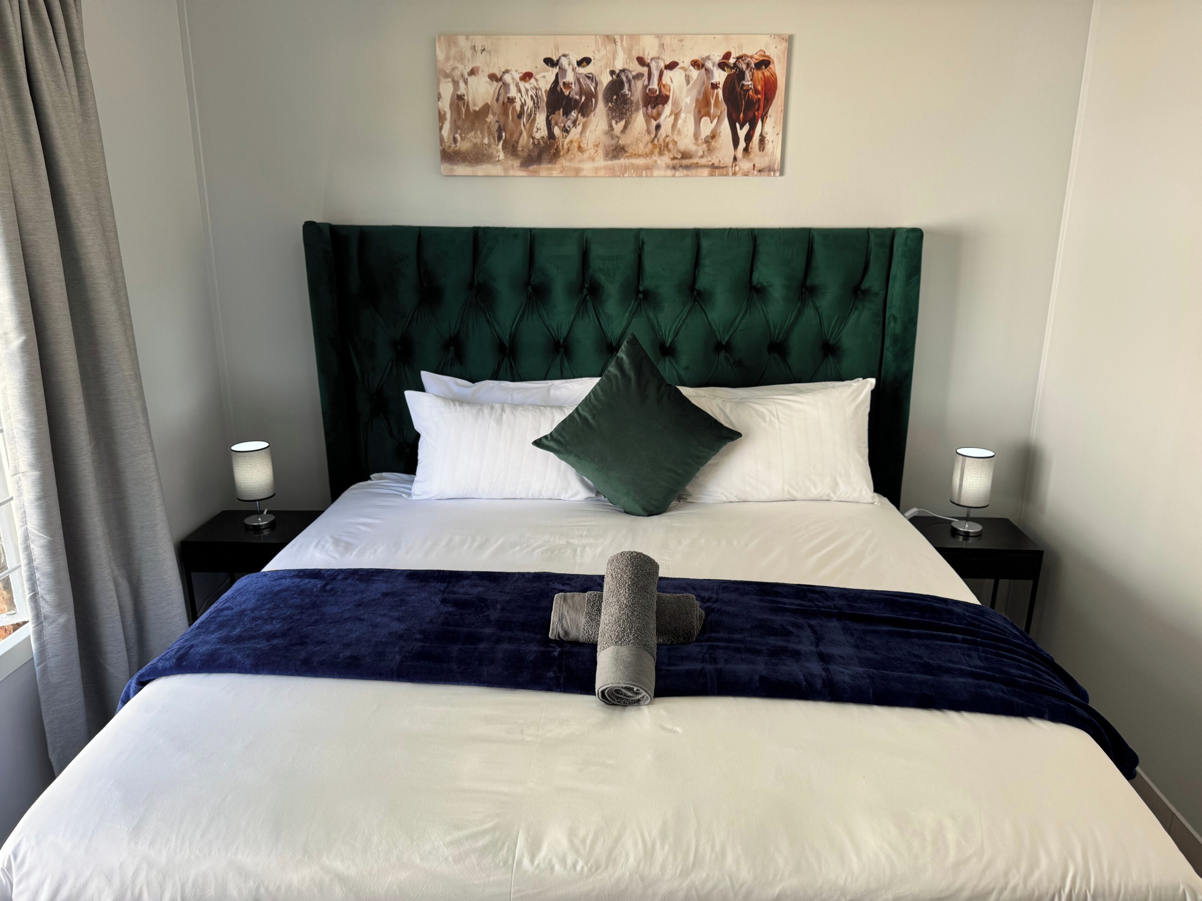 Una cama bien hecha, con un cabecero tapizado en verde, almohadas blancas y una manta azul oscuro. Hay una toalla en la cama y un cuadro de vacas en la pared sobre ella.