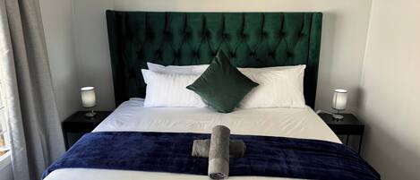 Una cama bien hecha, con un cabecero tapizado en verde, almohadas blancas y una manta azul oscuro. Hay una toalla en la cama y un cuadro de vacas en la pared sobre ella.